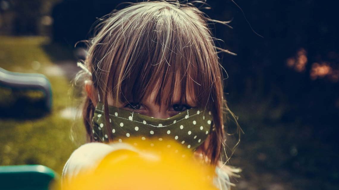 Kind mit Maske hält der Kamera einen Lolli hin.