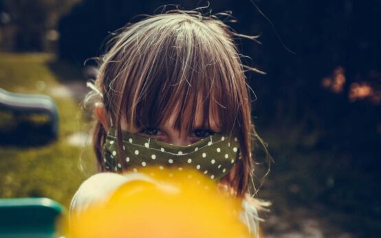 Kind mit Maske hält der Kamera einen Lolli hin.