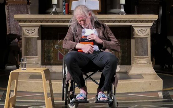 Auf der Bühne sitzt ein alter Mann im Rollstuhl. Lange, weiße Haare, ungepflegter Bademantel, nackte Füße.