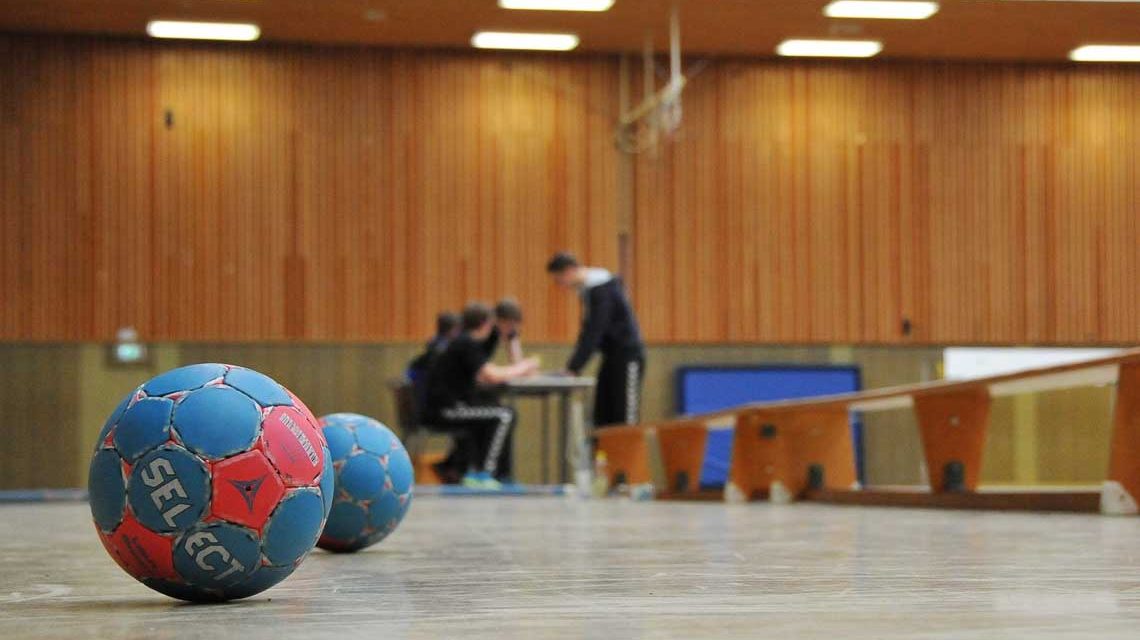 Handball im Vordergrund, hinten Vereinsmitglieder an einem Tisch stehend in einer Sporthalle. Energiekostenzuschüsse helfen, dass das Licht nicht aus geht.