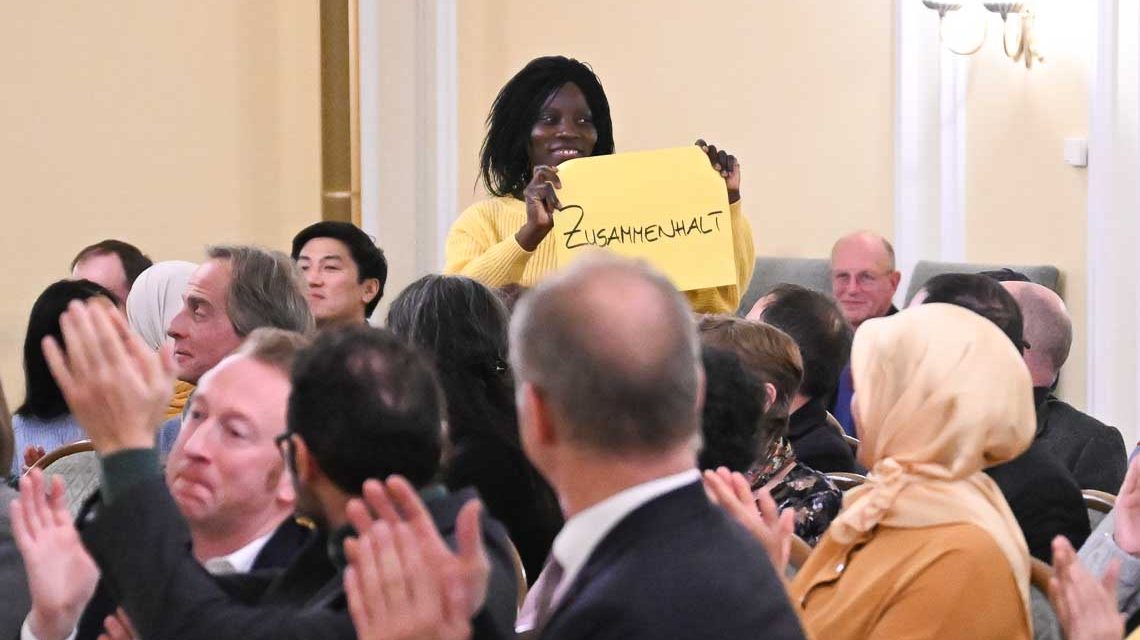 Frau steht auf im Festsaal des Rathauses und hält ein Plakat mit hoch. Darauf steht „Zusammenhalt"