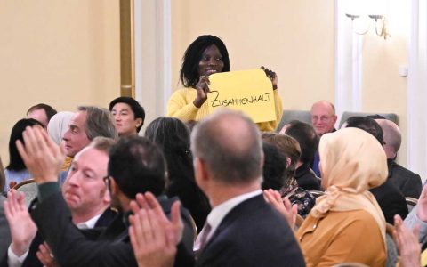 Frau steht auf im Festsaal des Rathauses und hält ein Plakat mit hoch. Darauf steht „Zusammenhalt"
