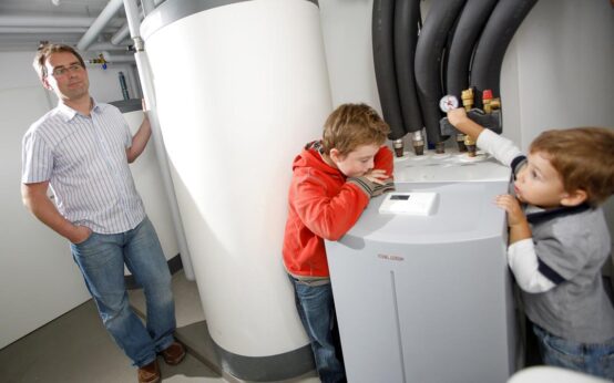 Wenn die Eltern auf die Wärmepumpe setzen freut sich der Nachwuchs: Um Bild steht lehnt der Vater an die neue Technik, während Kinder die Zähler ablesen.