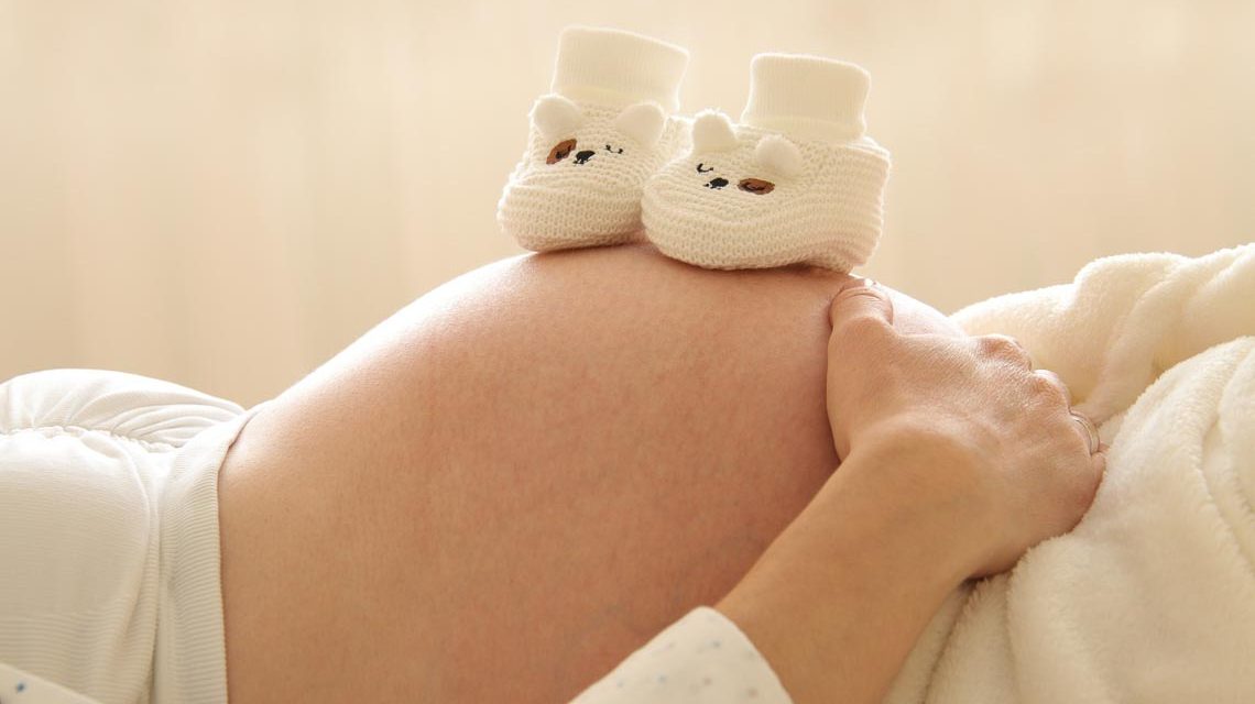 Schwangere Frau auf dem Rücken liegend. Auf dem Babybauch stehen Babyschuhe. Verständliche Medizin informiert schwangere Frauen und werdende Eltern