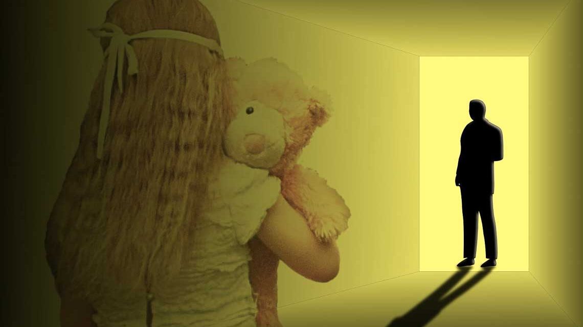 Mädchen mit Teddybär im Arm schaut zur Tür, wo ein Erwachsener steht.