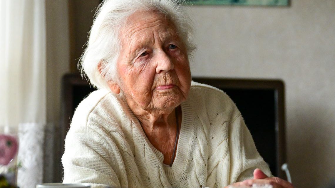 Gisela Voje, 103 sitzt in ihrem Wohnzimmer am Tisch.