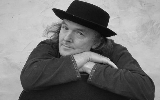 Gerhard Schöne kommt nach Wiesbaden. Im Bild sitzt er auf einem Stuhl und schaut verträumt in die Kamera.