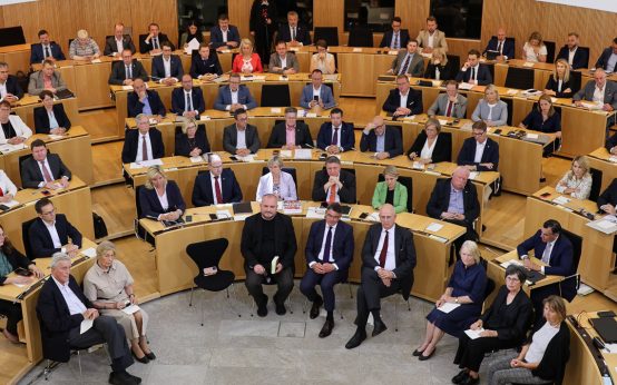 Landtagsabgeordnete sitzen im Plenum und hören der Lesung von Tim Pröse zu.