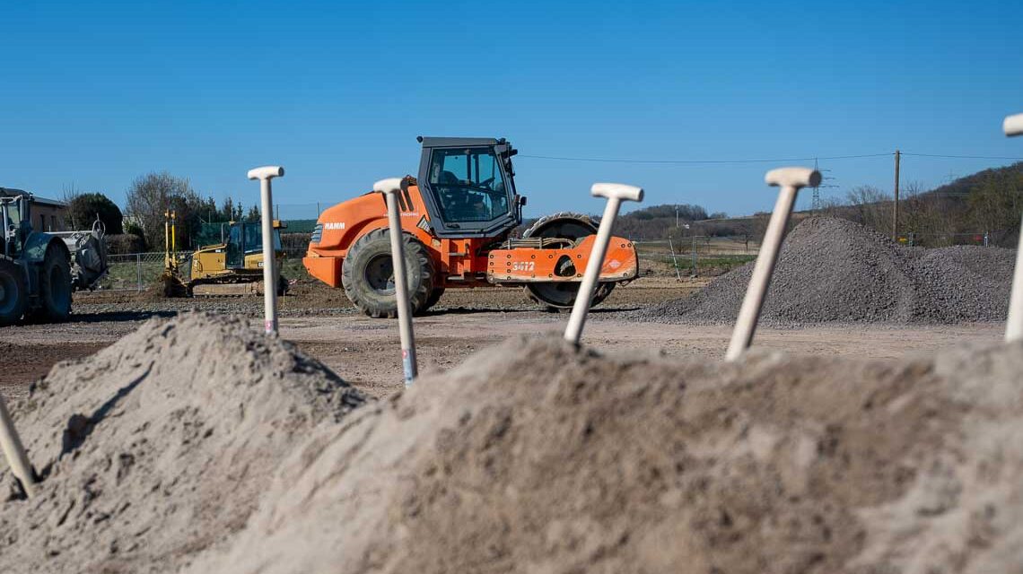 Spatenstich für neue HSK-Wohnungen, vier Spaten stecken in einem Sandhaufen, im Hintergrund ist ein Baufahrzeug.