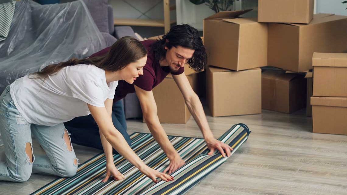 Frau und Mann rollen einen Teppich aus. Im Hintergrund stehen Kisten.