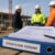 Personen auf einer Baustelle im Gespräch. Davor ein Tisch mit einem Ordner „Öffentliche vergabe"